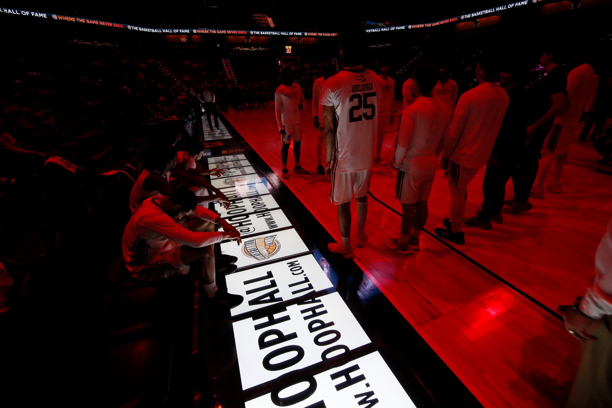 Players on the bench beside glowing courtside LED floor panels showing Basketball Hall of Fame ads before tip-off