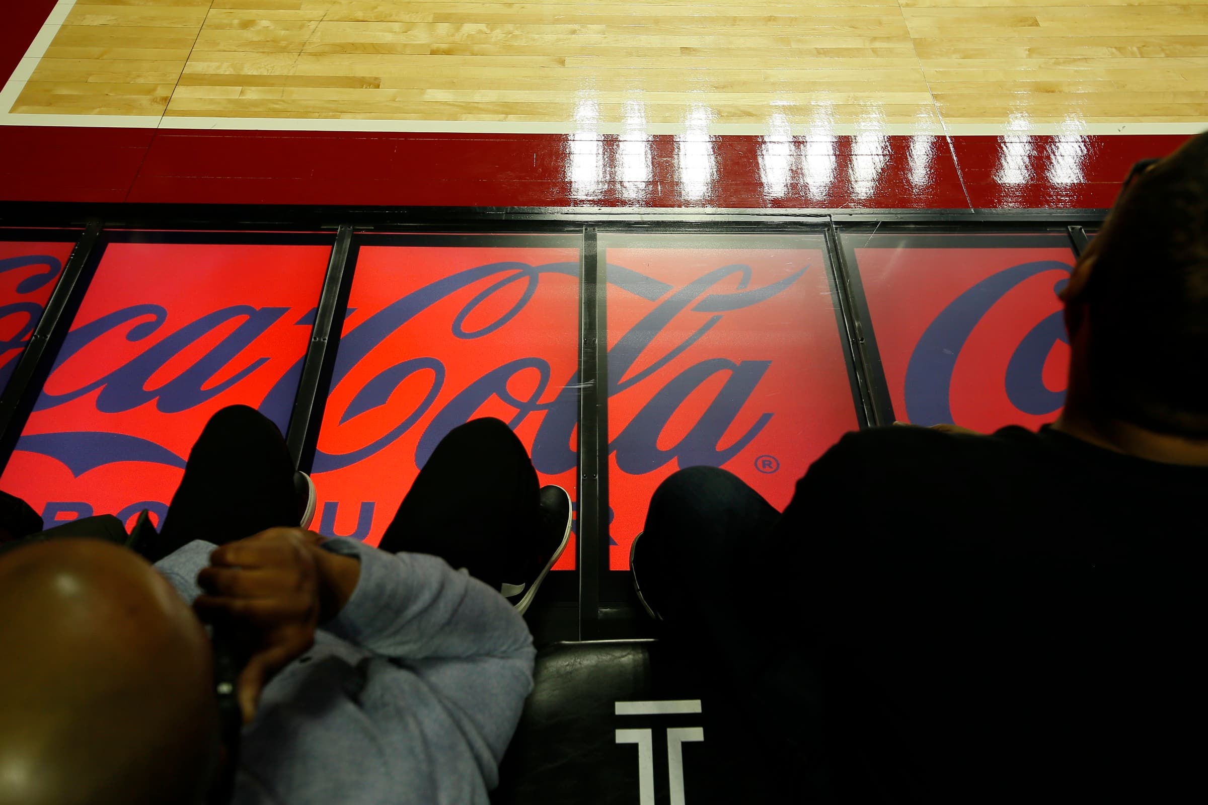 Courtside fans' feet resting beside a row of floor-level panels showing a Coca-Cola ad along the court's edge